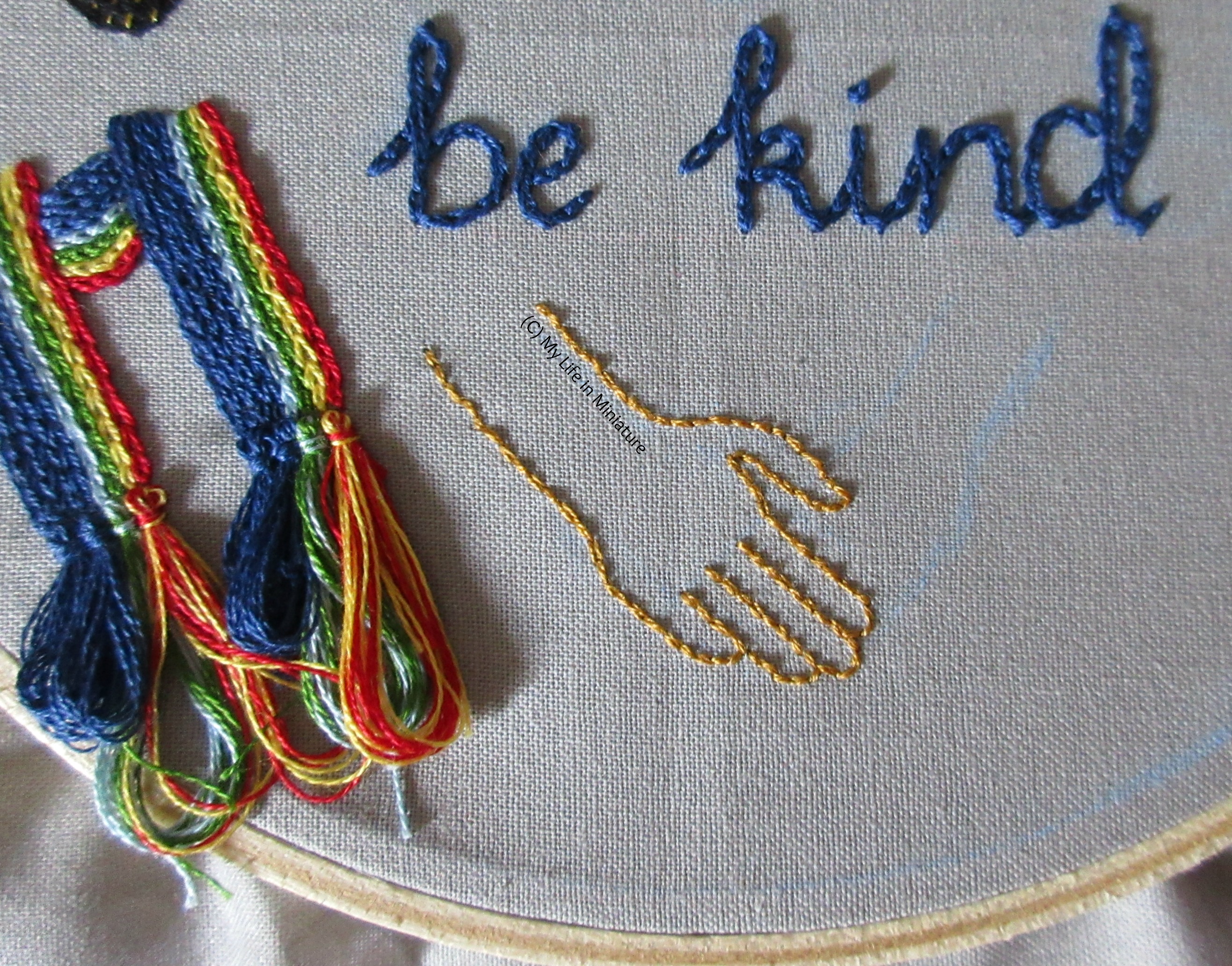 The bottom of the grey hoop is visible, with the scarf on the left. The outline of a hand reaching out as if to shake hands is stitched in gold. 