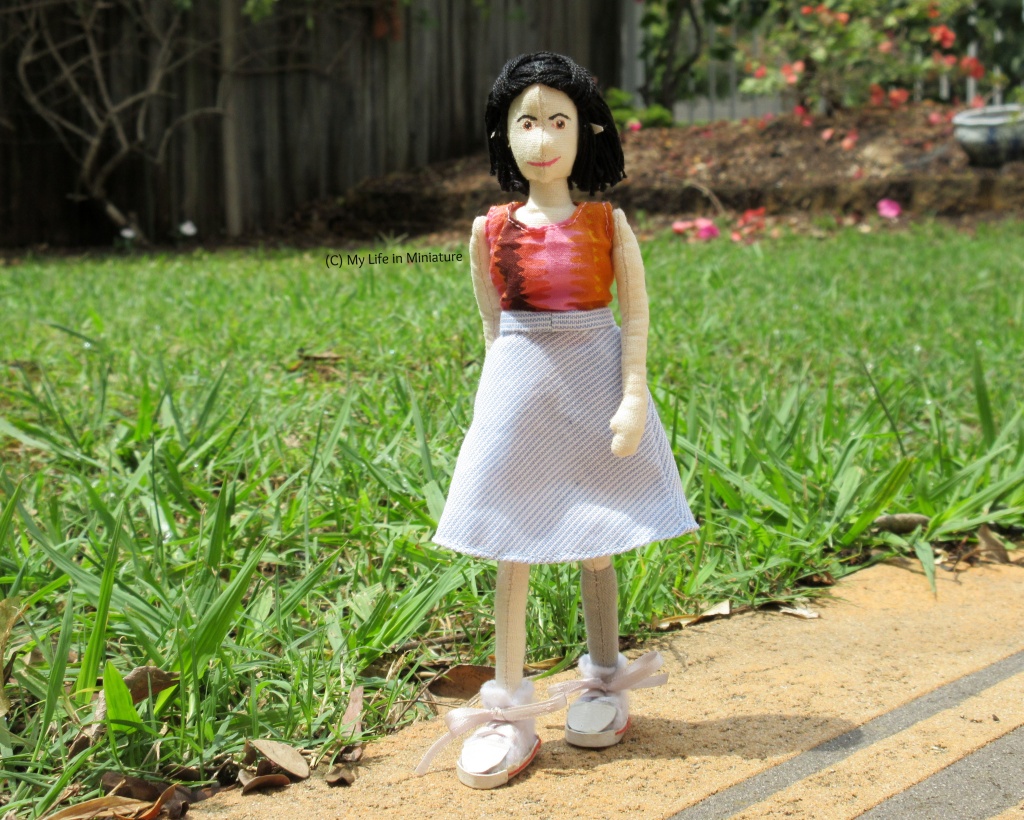 Tiffany walks along a garden path towards the camera. She wears a blue-and-white striped skirt and a pink-and-orange top.