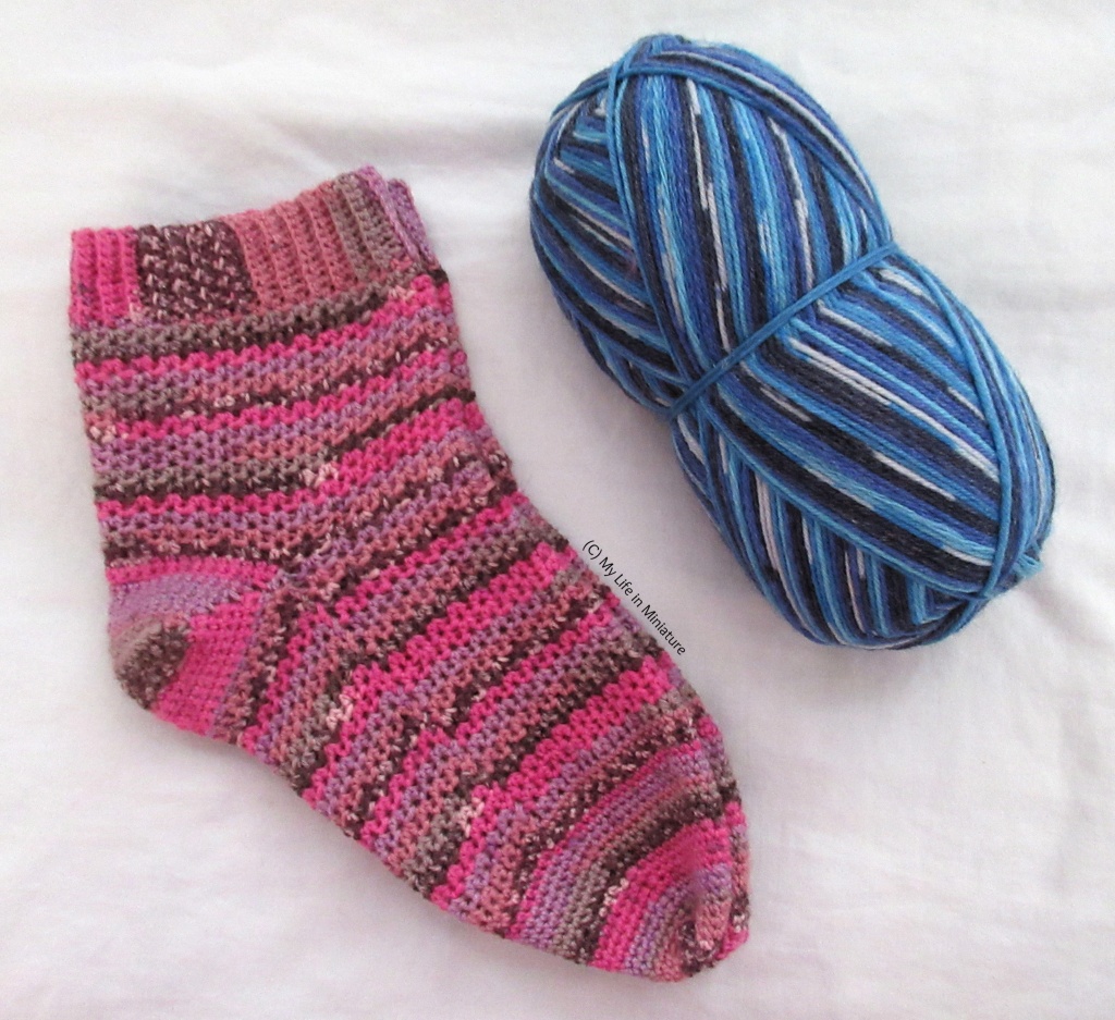 The socks, stacked on top of each other, lie on the grey fabric background next to a large ball of blue yarn. The blue yarn is striped in shades of white and blue like the pink yarn.