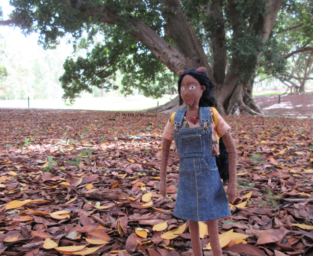 Petra walks towards the camera , looking slightly to the left. Behind her is an enormous tree, and on the ground is a thick groundcover of brown and yellow leaves. 
