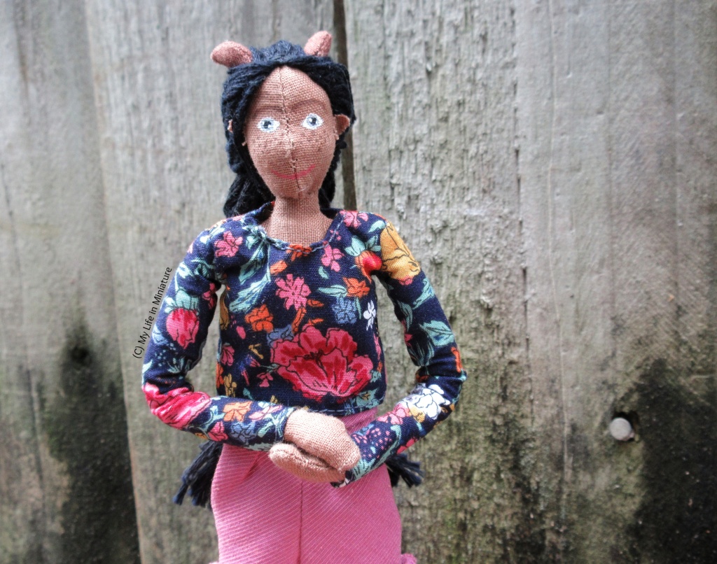 Petra wears her navy floral long-sleeved top with pink corduroy shorts. She is outside, against a wooden fence. Her hands are clasped in front of her and she smiles at the camera.