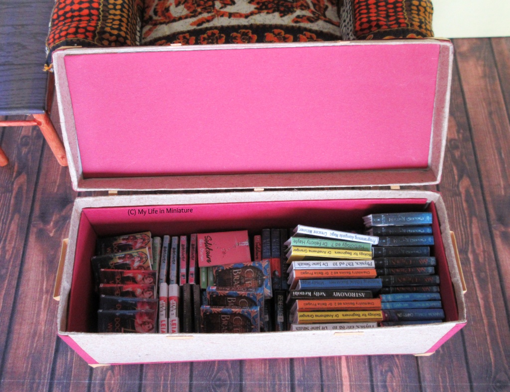 View of the inside of the trunk in the upstairs room of the Palace Library. The inside of the trunk is in the same dark red as the trim, and it's filled with miniature books. Also visible on the outside are three gold hinges on the back, and two gold handles on either side of the trunk.