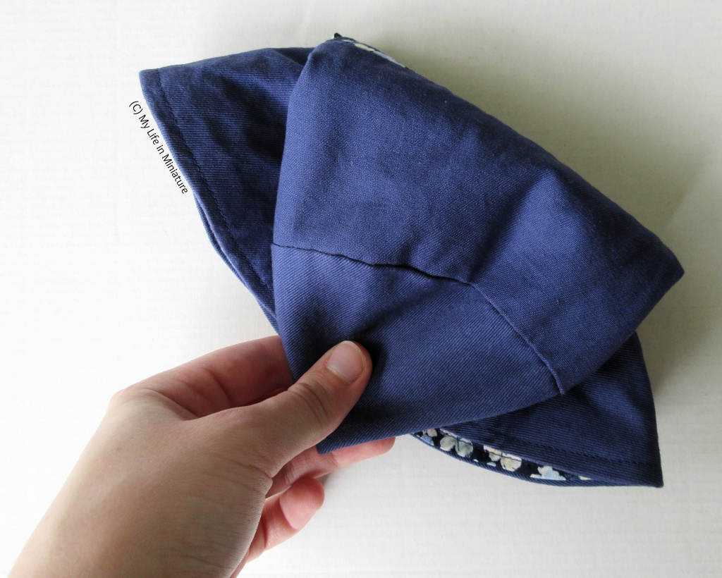 The navy blue hat sits on a white background. It is folded up small, and the author's white hand is holding it. 