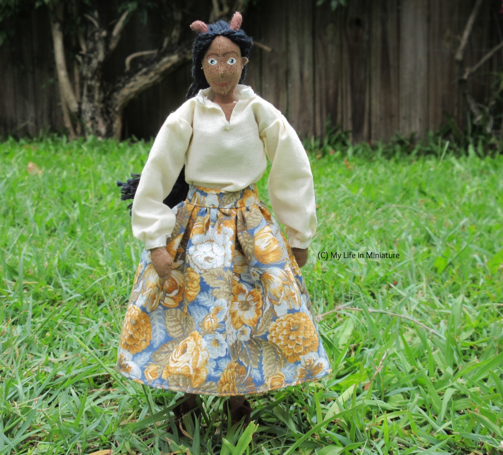 Petra stands on grass, with a wooden fence and a bougainvillea hedge in the background. She wears a white 'pirate shirt' with a voluminous floral skirt, in shades of white, blue, and orange. She has just come to a standstill facing the camera after twirling on the spot; her face is flushed, her long plait swings. 