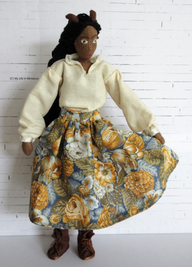 Petra stands in front of a white brick background. She wears a white 'pirate shirt' with a voluminous skirt in a blue, orange, and white floral pattern and brown boots. She is holding the skirt out, making the pattern visible and smoothing out the gathers. Petra looks at her hand as it does this.