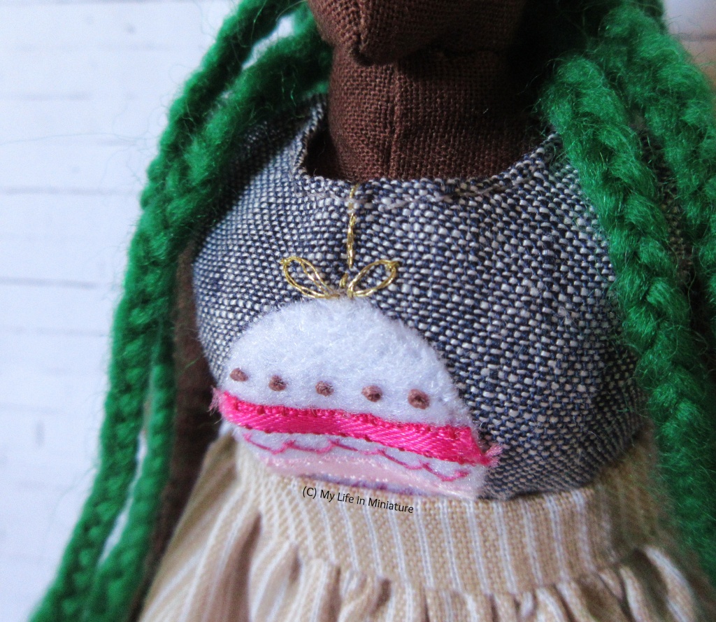 Close-up of the bauble on Hazel's shirt, and particularly the gold embroidered hanging thread above the bauble. Her braids and skirt waistband are also visible in the shot, as is a white brick background.