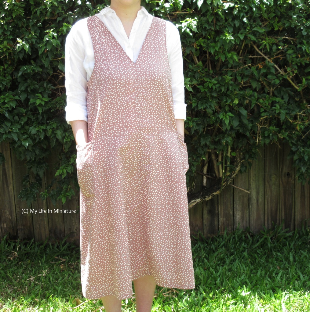 The author stands in front of a hedge. She wears the same white shirt/pink shift dress combo as the previous image. Her hands are in the two patch pockets on the sides of the dress, and it is clearer now that the dress is midi-length. 