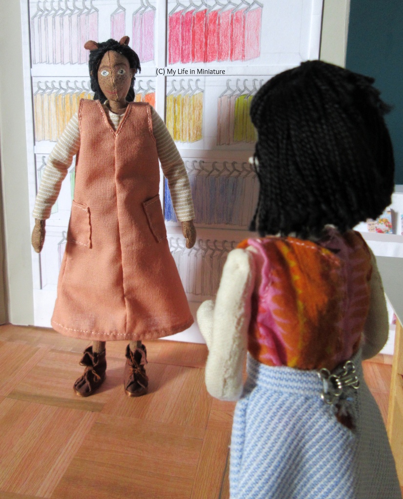 Petra stands in front of the fat quarters in Needle & Thread, wearing the peach dress over her beige shirt and with her brown boots. The peach dress is about midi-length, V-necked with two patch pockets. She faces the camera and Tiffany, who is just in front of the camera with her back to it. Tiffany is excited to see Petra in the dress, though her expression isn't visible.