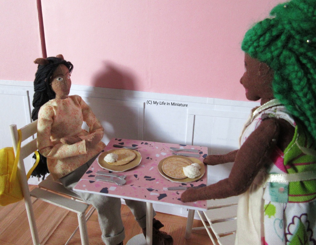 Hazel moves to sit at one of Fierro's' tables across from Petra, who is already sitting. Petra wears a beige long-sleeved peplum shirt with grey pants, and is smiling at Hazel. In front of each seat is a plate of pancakes with ice cream, and a knife, fork, and spoon. Petra's yellow backpack hangs over the back of the white chair.