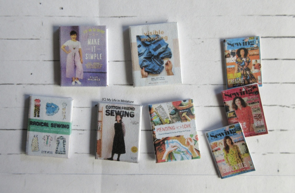 Five miniature craft books and three miniature sewing magazines are laid out flat on a white brick background. The five books are various titles about sewing clothes and mending fabrics. The three magazines are recent issues of 'Simply Sewing'. 