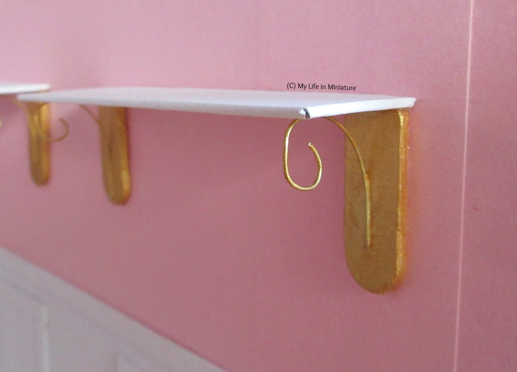 Close-up of one of the brackets for the shelves. It is gold; it has a flat wooden bit against the wall, with a gold wire attached that curves up to support the shelf. The shelf itself is white. 