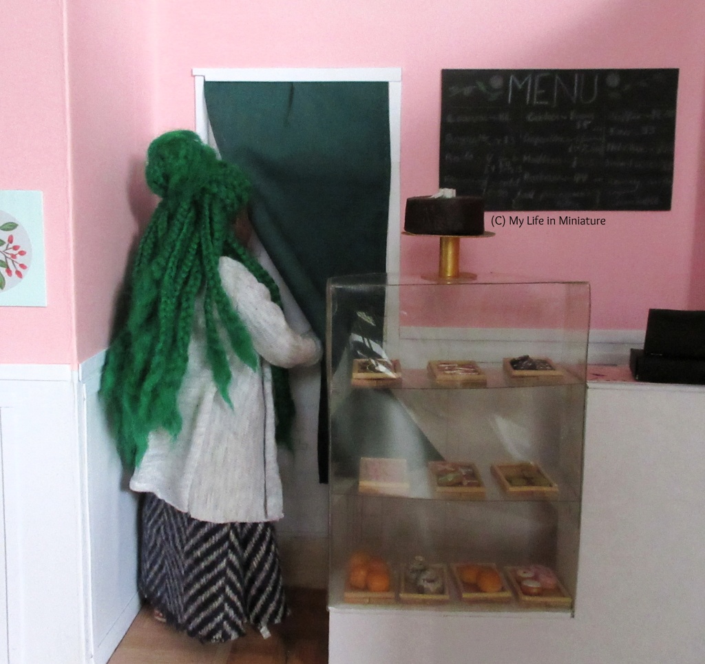 Hazel pushes aside the dark green curtain obscuring a doorway beside the menu and behind the counter. Her green braids fall down her back.