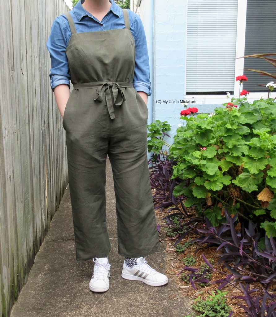 The author stands on a garden path beside a garden bed. She wears a pair of green linen dungarees with white shoes and a denim blue shirt. The dungarees have a tie at the waist, and a square neckline. Her hands are in her pockets. 