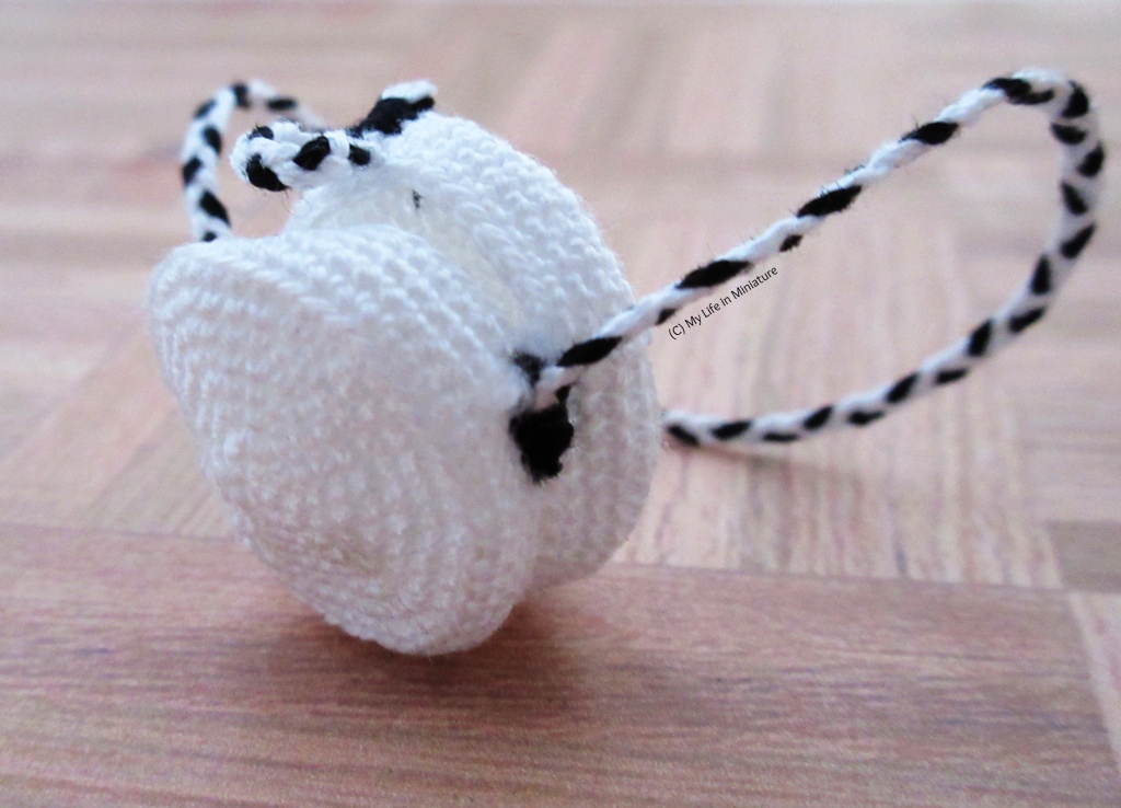 Close-up shot of the crocheted bag on a pale wood surface. The bag is at an angle, so one can see the two sides sewn together, as well as one side of the black-and-white strap where it attaches near the opening. A small black-and-white loop is stitched onto the top of the bag as a closure.