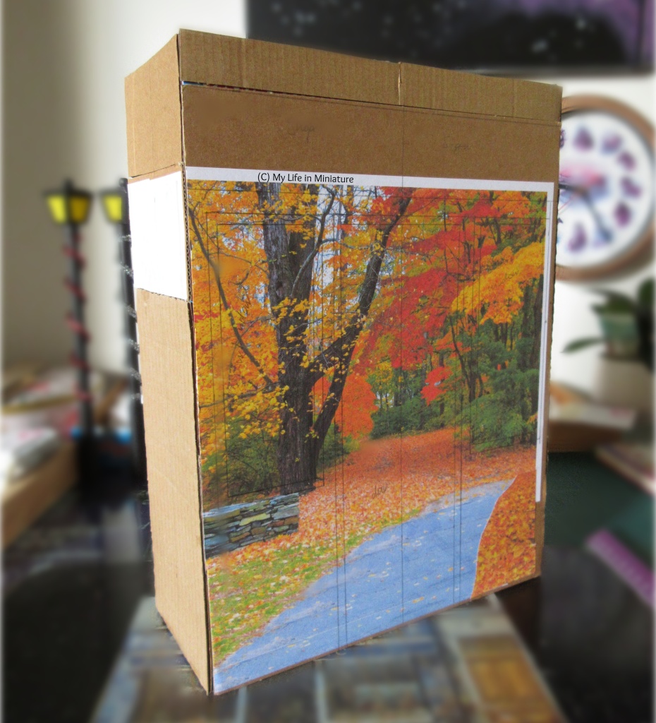 A rectangular box standing vertically sits on a desk, angled so that the side and the front are both visible. It is about the same size as a cereal box, and has cardboard glued over the entire outside. On the cardboard are pencil lines, marking features of the facade. The background is blurred.