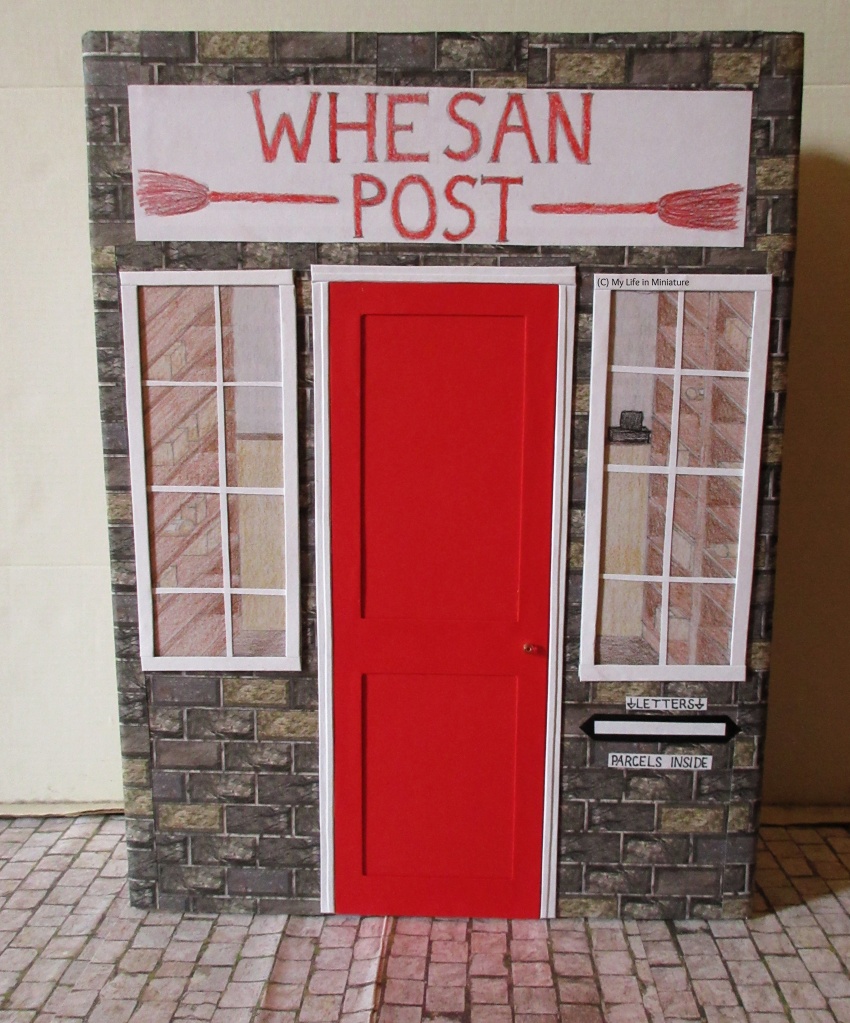 The completed storefront stands on cobblestones, against a white background. The door has a doorframe and a handle, the windows have frames and glass panes, and there is a slot under the right-hand window for posting letters. 