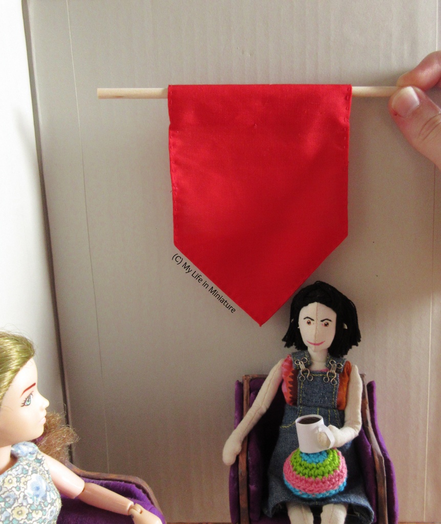 A hand holds a wooden dowel, from which hangs a red banner, against the wall behind Tiffany. Tiff is still sitting in the armchair, drink in hand. The banner is rectangular with a pointed bottom edge. 