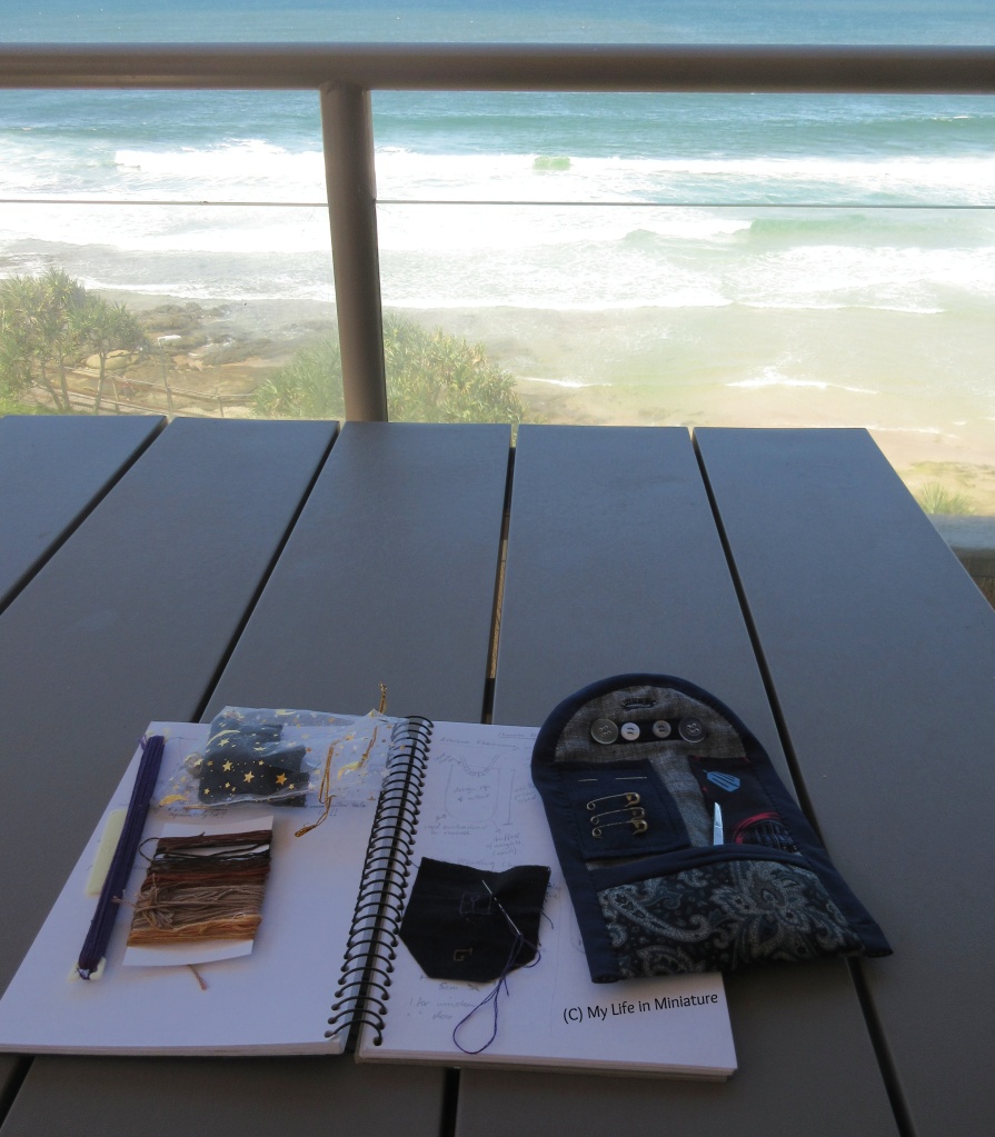 A table sits at the edge of a balcony, overlooking the sea. On the table is an open sketchbook, with small pieces of fabric, threads, and the travel sewing kit on top. One of the pieces of fabric has a needle in, mid-stitch.