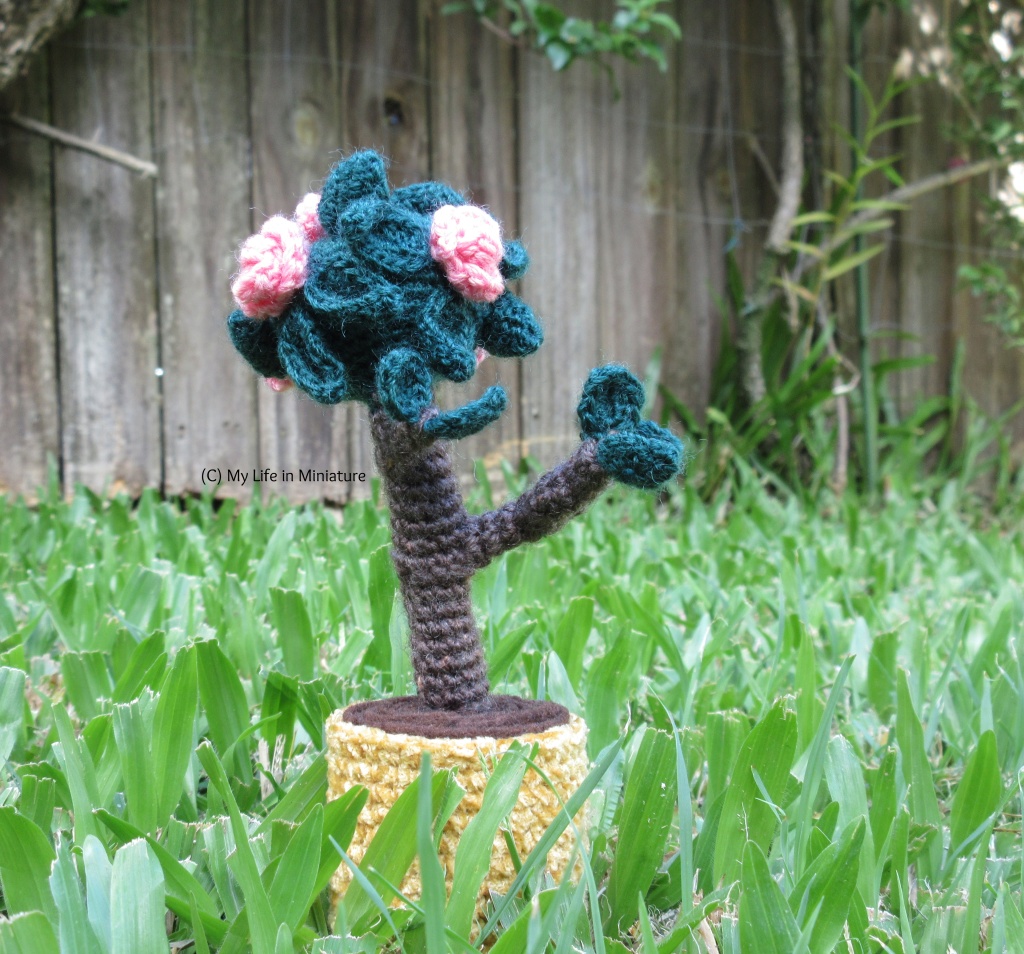 A small crocheted tree in a yellow pot stands on grass, with a wooden fence and a larger tree in the background. The tree has branches with leaves budding on the ends, and pink flowers blooming on the canopy. 